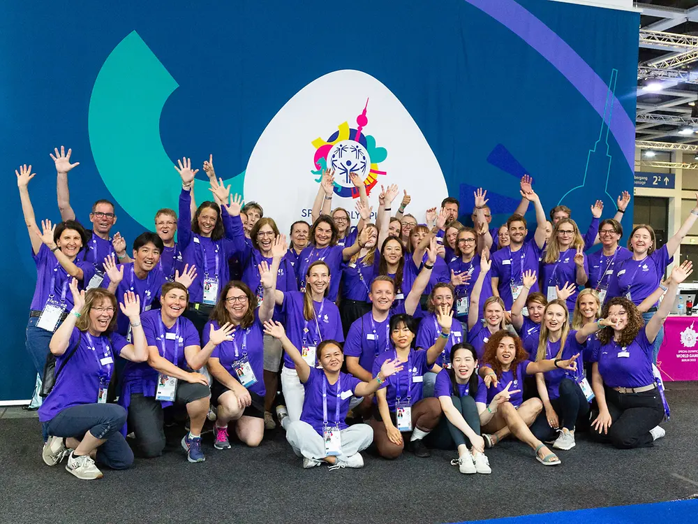 A group of volunteers in purple shirts posing together and cheering at a Special Olympics event.