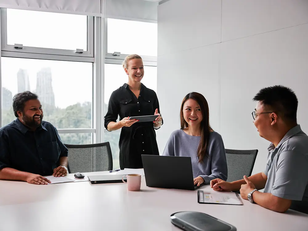 A diverse group of professionals in a modern meeting room, collaborating around a table with laptops and documents. The scene reflects Henkel’s commitment to gender parity and diversity in leadership, showcasing an inclusive work environment where different perspectives contribute to decision-making.