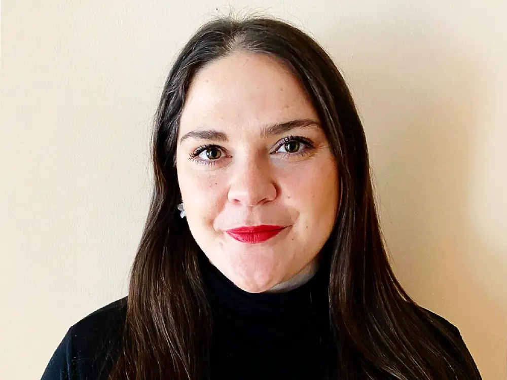 
Recruitment partner from North America wearing a black top, standing against a light beige background, sharing advice on how to craft a successful career. 