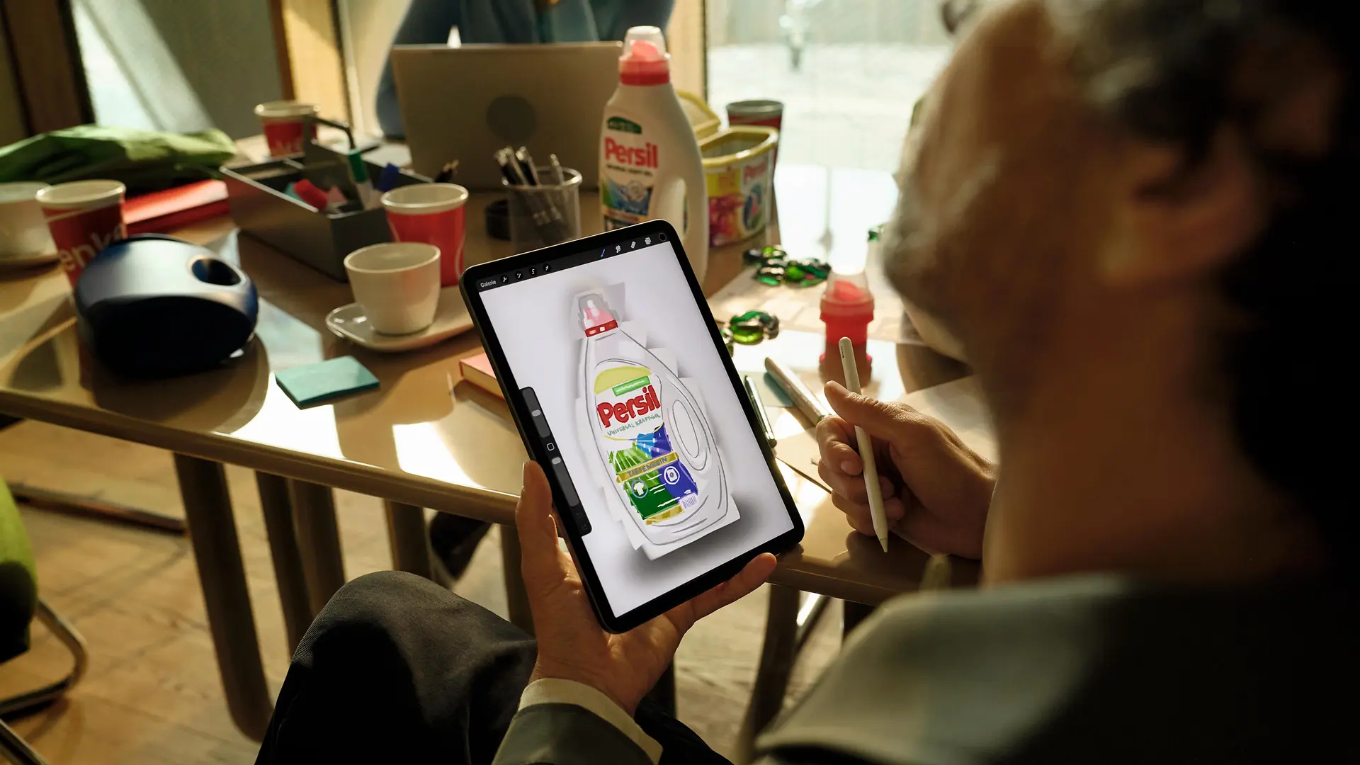 A person holding a tablet displaying a detergent bottle in a meeting room with other detergent products on the table.